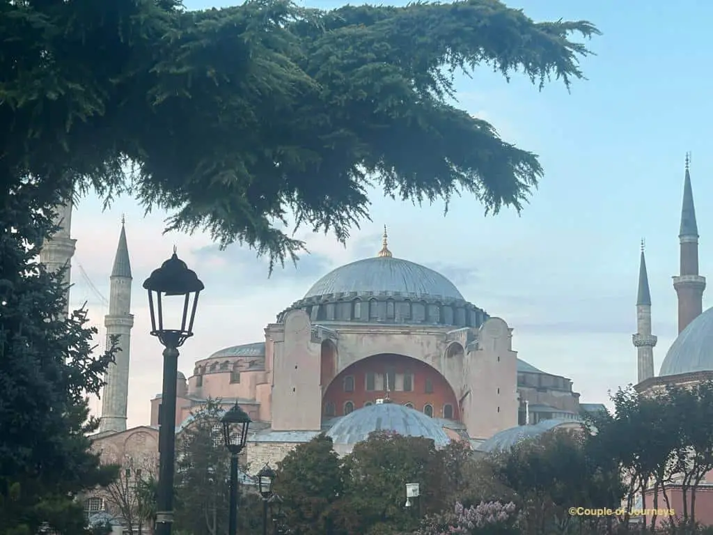 A serene view of the Hagia Sophia in Istanbul, framed by lush greenery and a vintage lamp post, with its grand domes and towering minarets standing out against the soft blue sky—an enchanting moment perfect for Istanbul Instagram captions and quotes about history and beauty