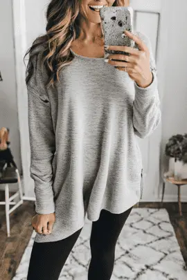  A woman in a loose gray sweater and black leggings, taking a mirror selfie in a cozy indoor setting, demonstrating a comfortable and chic Munich travel outfit for lounging or casual outings.