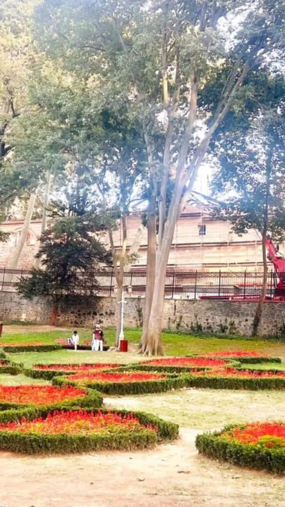 A tranquil park scene in Istanbul featuring tall trees, manicured gardens with red flowers forming patterns, and a couple of people sitting on a bench near a historical stone wall, captured for an Istanbul Instagram story.