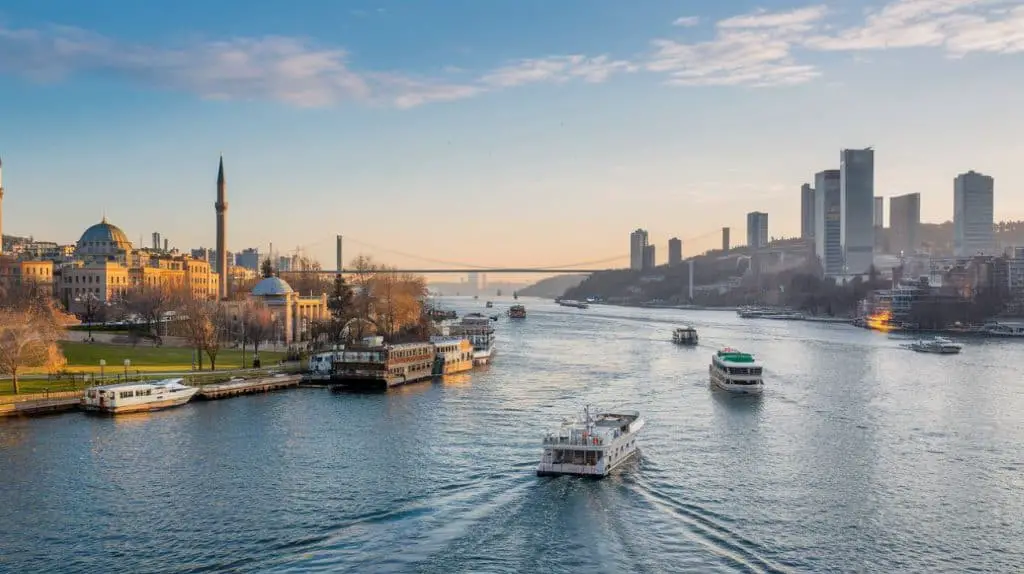 A vibrant aerial view of the Bosphorus Strait in Istanbul under a bright blue sky, with the iconic suspension bridge connecting two continents, boats navigating the shimmering water, and lush parks and buildings lining the shores—perfect for Istanbul Instagram captions and quotes celebrating the beauty of this city.