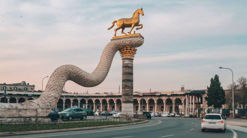  A historical monument featuring a striking golden horse atop a column shaped like a serpent, with arched structures and a clear sky in the background.