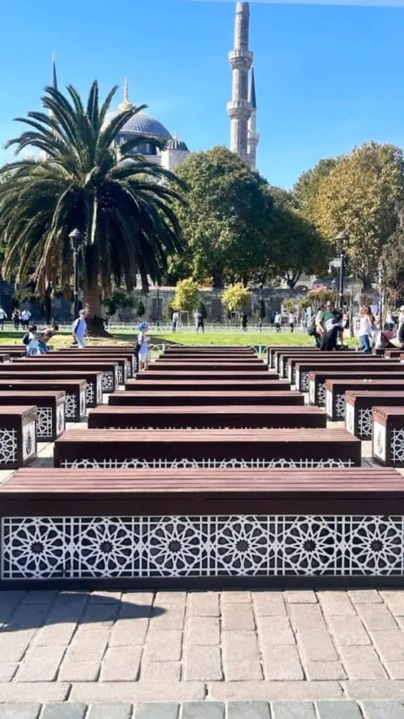 Rows of intricately designed wooden benches leading to a scenic view of a mosque surrounded by palm trees and greenery in Istanbul, showcased in an Istanbul Instagram story.