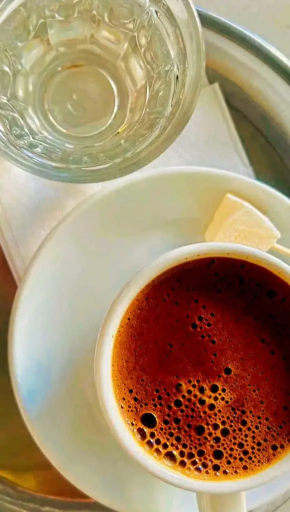 A traditional Turkish coffee cup on a saucer, filled with dark, frothy coffee, placed alongside a glass of water and a small piece of Turkish delight on a tray, presented in an Istanbul Instagram story.