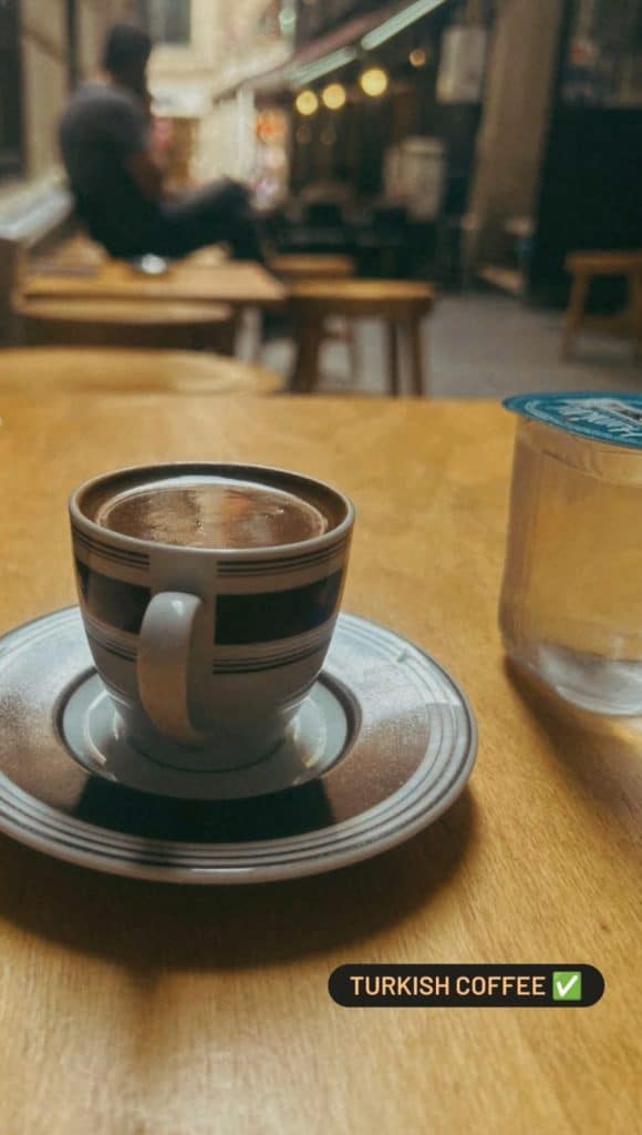 A cozy café scene with a cup of Turkish coffee served on a patterned saucer, set on a wooden table with a blurred background of people sitting in an alleyway, ideal for an Istanbul Instagram story.