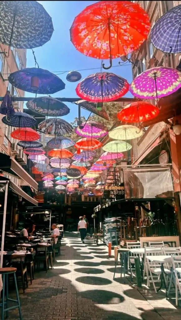 A vibrant alley in Istanbul adorned with colorful umbrellas hanging overhead, casting playful shadows on the ground, with outdoor seating and a lively café atmosphere, perfect for an Istanbul Instagram story.