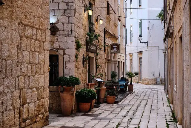 A charming cobblestone alley in Croatia lined with rustic stone buildings adorned with plants in clay pots, with small shop signs and hanging lights adding a quaint atmosphere.