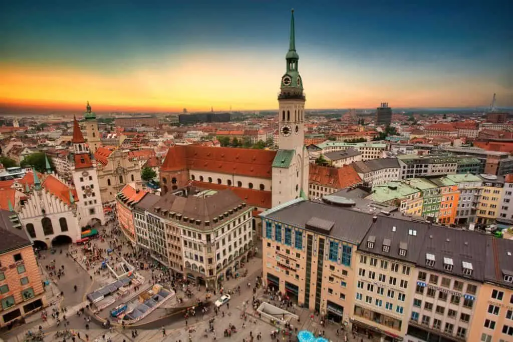 Aerial view of Munich’s Marienplatz at sunset, featuring St. Peter’s Church (Alter Peter) with its striking clock tower and red rooftops. The bustling square is filled with people and surrounded by colorful historic buildings. A must-visit location in any Munich Itinerary 3 Days