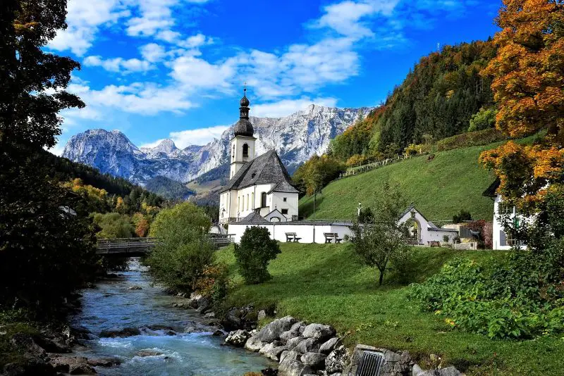 A picturesque riverside scene in Berchtesgaden, Germany, featuring a charming white church and the stunning backdrop of the Alps—one of the most romantic honeymoon destinations in Germany for couples seeking tranquilityphotos