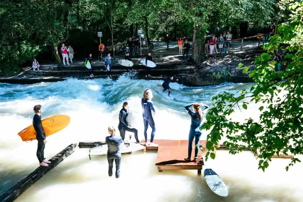 A group of surfers in wetsuits waiting their turn at the famous Eisbachwelle in Munich, a unique man-made wave in the middle of the city. A surfer is seen riding the powerful river wave while spectators watch from the banks, making this urban surfing spot one of the most exciting and unexpected "Things to do in Munich."