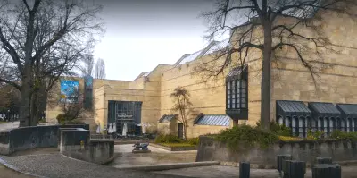 Exterior view of the Neue Pinakothek in Munich, featuring its modern, angular stone façade with large windows and minimalistic design. As one of the top cultural attractions among things to do in Munich, the museum is surrounded by leafless trees and a cobblestone pathway, with outdoor seating near the entrance, perfect for art enthusiasts exploring 19th-century masterpieces.