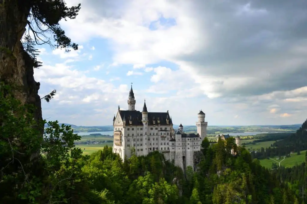 Neuschwanstein Castle, a fairytale-like fortress perched atop a lush, green hill in Bavaria, Germany, with rolling countryside and a dramatic cloudy sky in the background. This iconic castle, one of the most famous landmarks near Munich, is a must-visit for travelers exploring the best "Things to do in Munich." Its elegant white facade, towering spires, and picturesque surroundings make it a dreamlike destination straight out of a storybook.