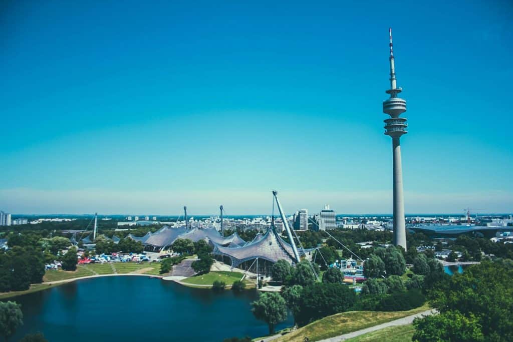 A panoramic view of Munich’s Olympic Park, featuring the futuristic tent-like roofs of the Olympic Stadium, a serene lake, and the towering Olympiaturm against a clear blue sky. This historic site from the 1972 Olympics is now a popular destination for sports, concerts, and city views, making it a top highlight in "Things to do in Munich."