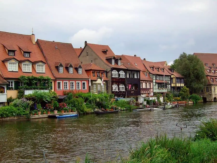  A picturesque row of colorful half-timbered houses with red-tiled roofs lines the riverbank in Bamberg, Germany. The lush greenery and potted plants in front of the homes add to the charm, while boats are moored along the calm water. A must-visit spot for those exploring Germany travel destinations.