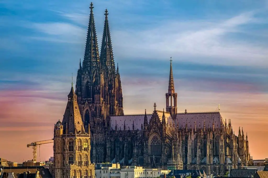 The stunning Cologne Cathedral dominates the skyline, with its intricate Gothic spires towering over the city. The golden glow of sunset bathes the historic architecture, with the foreground featuring a mix of modern and historic buildings. A breathtaking highlight among Germany travel destinations.