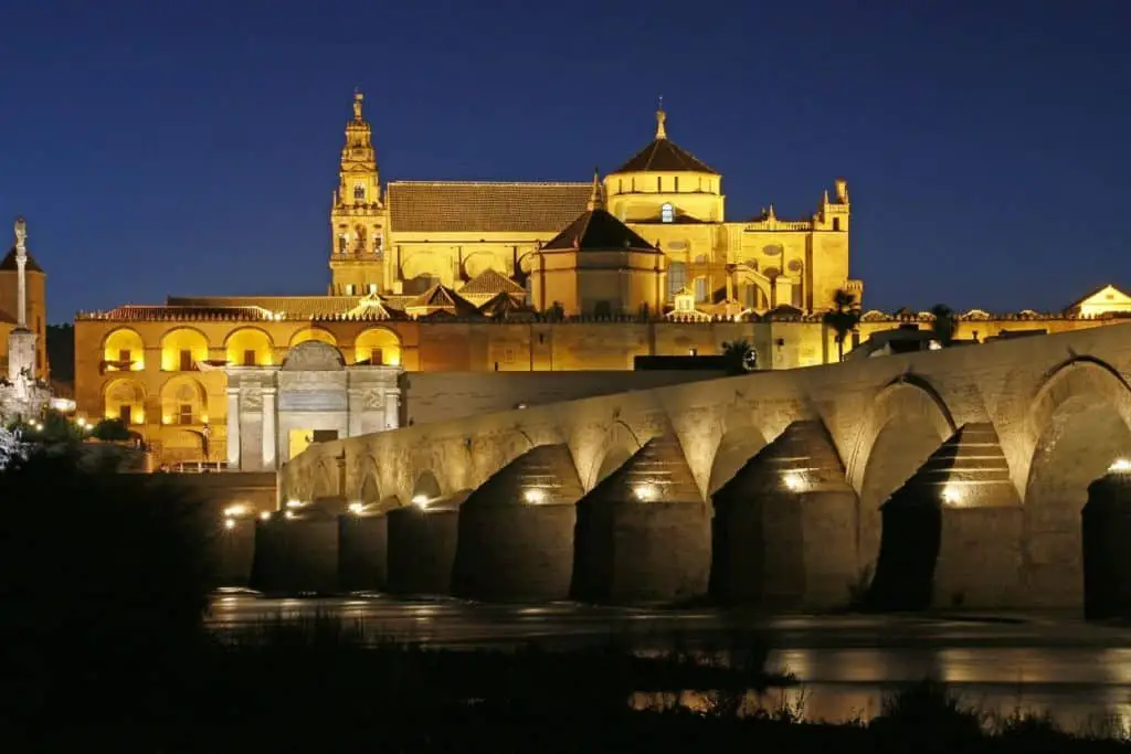  A stunning nighttime view of the illuminated Mosque-Cathedral of Córdoba and the historic Roman Bridge, making it a cultural gem among Cheap Travel Destinations in Europe Summer.