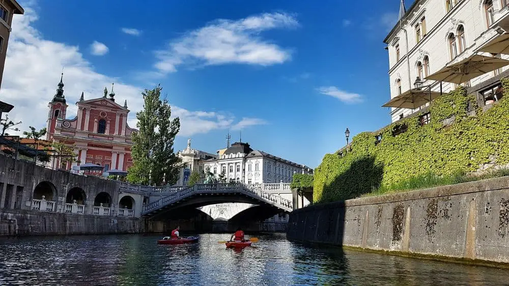 A scenic canal view with kayakers, a historic bridge, and the iconic Franciscan Church, making Ljubljana a top choice for Cheap Travel Destinations in Europe Summer.