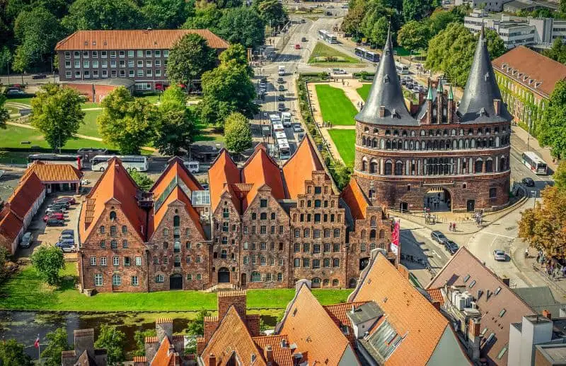 An aerial view of Lübeck, Germany, featuring the iconic Holstentor, a medieval red-brick gate with twin round towers. Surrounding the landmark are historic gabled buildings with red rooftops, lush green parks, and bustling city streets. A top historical site in Germany travel destinations.