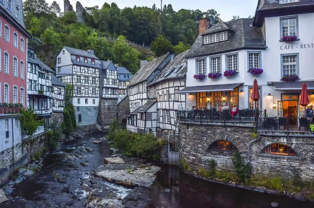 A charming European village with half-timbered houses lining a narrow, rocky river in Monschau, Germany. A cozy café with an outdoor terrace, adorned with flower boxes and red umbrellas, overlooks the water, while the ruins of an old castle sit atop a lush green hill in the background. A fairytale-like setting in Germany travel destinations