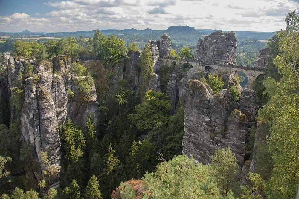 Towering sandstone rock formations covered in greenery create a dramatic landscape in Saxon Switzerland National Park, Germany. A historic stone bridge spans between the rugged cliffs, with a breathtaking backdrop of rolling hills and forests. An unmissable adventure spot in Germany travel destinations.