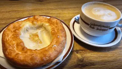 A traditional Bavarian Schmalznudel, golden and crispy with a slightly hollow center, served on a plate alongside a frothy cup of coffee from Café Frischhut in Munich. Enjoying local pastries like this is one of the delicious things to do in Munich, offering a flavorful glimpse into the city’s rich café culture.