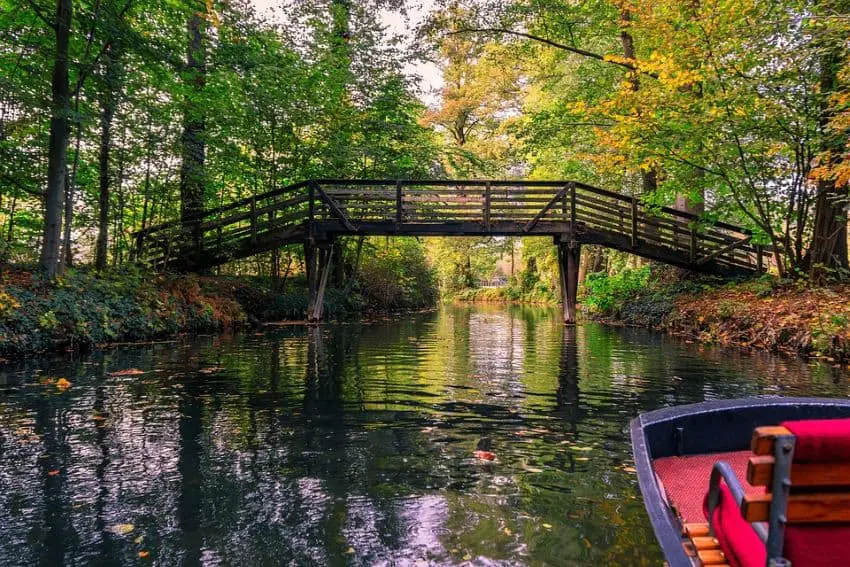  A serene waterway surrounded by lush green trees with hints of autumn foliage. A rustic wooden bridge arches over the canal, while a small boat with red seating drifts in the calm, reflective water. This peaceful nature escape is a hidden gem among Germany travel destinations.