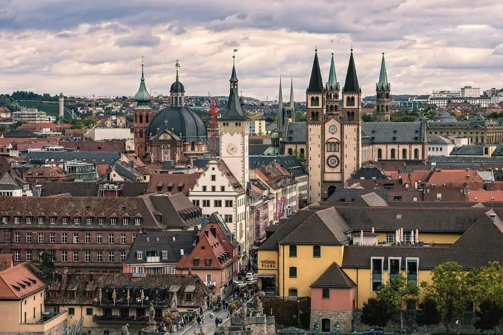 A panoramic view of Würzburg, Germany, showcasing its historic skyline with the twin spires of St. Kilian Cathedral and the domed church standing prominently. The cityscape is filled with red-roofed buildings, winding streets, and a lively urban atmosphere. This historic city is a top pick for Germany travel destinations