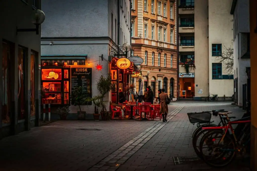 Alt text: A charming European street in Munich at dusk, featuring a cozy kiosk and café with warm glowing lights. Red chairs are set outside, where a few people in winter coats are chatting, adding to the inviting atmosphere. Historic buildings with ornate facades line the background, while bicycles are parked in the foreground—capturing the essence of a perfect stop on a 3 days itinerary Munich adventure.