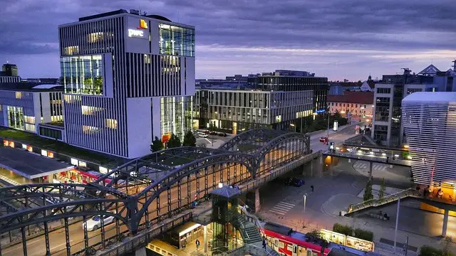 Evening view of the NS-Dokumentationszentrum in Munich, a modern building with sleek, white architecture illuminated by city lights. The surrounding area features contemporary office buildings and a glass-covered pedestrian bridge over a bustling street with trams. As one of the educational highlights among things to do in Munich, this site offers historical insights into the city's past, set against a dramatic, cloudy sky.