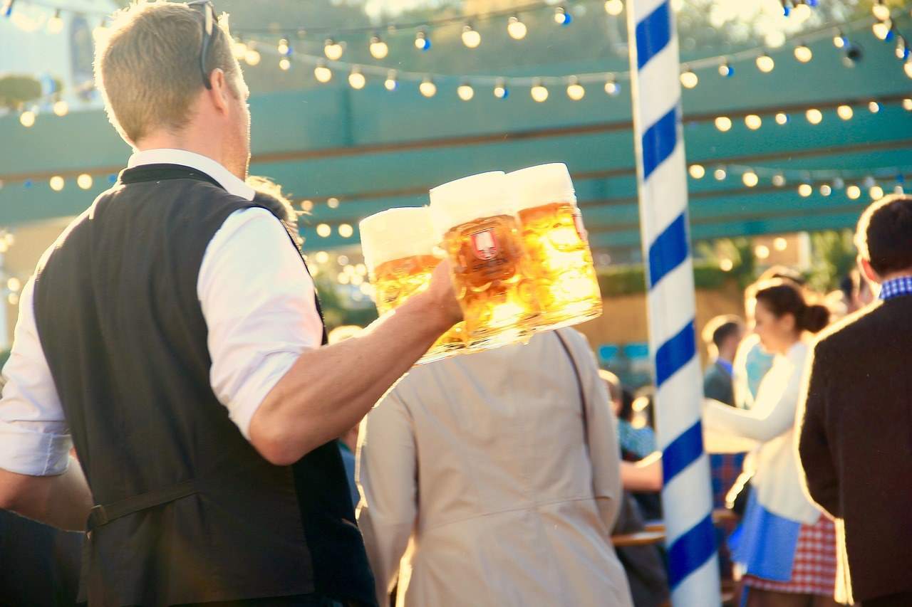 A man wearing a white shirt and black vest carries several large beer steins filled with golden beer, topped with frothy foam, at an Oktoberfest in Germany. String lights hang overhead, and people dressed in traditional attire mingle in the background. This lively scene captures the festive spirit of Oktoberfest, one of the top things to do in Munich.