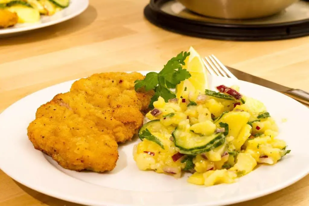 A delicious plate of traditional German food featuring a crispy, golden-brown schnitzel served with a side of vibrant potato salad mixed with cucumbers, red onions, and fresh herbs. A lemon wedge and a sprig of parsley add a fresh garnish. Enjoying authentic dishes like this is one of the top things to do in Munich for food lovers exploring Bavarian cuisine.