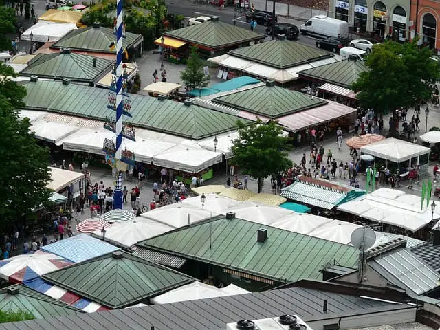 A bustling aerial view of Viktualienmarkt in Munich, showcasing numerous market stalls with green, white, and striped canopies, surrounded by crowds of people. The vibrant outdoor market features a traditional Bavarian maypole decorated with colorful symbols, adding to the lively atmosphere. As one of the must-visit spots among things to do in Munich, Viktualienmarkt offers fresh produce, local delicacies, and a rich cultural experience in the heart of the city.